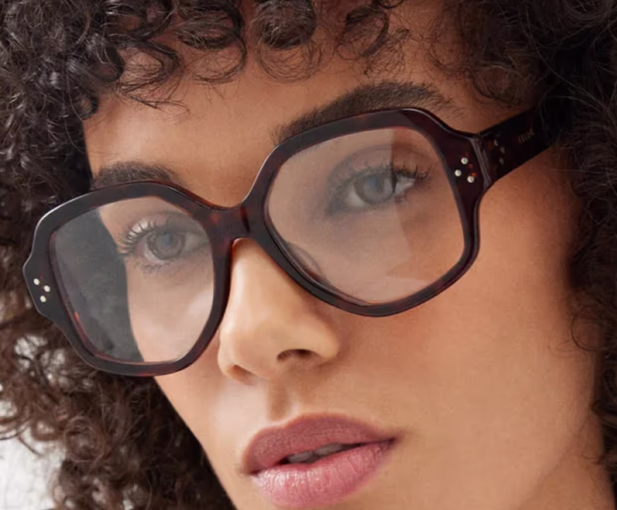 Close-up van een vrouw met krullend haar die oversized, vierkante bril met donker montuur draagt.
